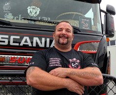 Ein Mann trgt ein schwarzes AST Fan Shirt aus Baumwolle/Viskose mit A-S-C Anker-Logo und "TRUCKING. FOREVER." Aufdruck. LKW im Hintergrund.