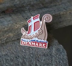Metall-Pin in Form eines Wikingerschiffs mit rot-weiem Segel und blauen Wellen. Schriftzug DENMARK.