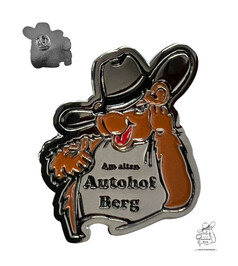 Metall-Anstecknadel in Form eines braunen Cartoon-Bren mit grauem Shirt und der Aufschrift "Am alten Autohof Berg".